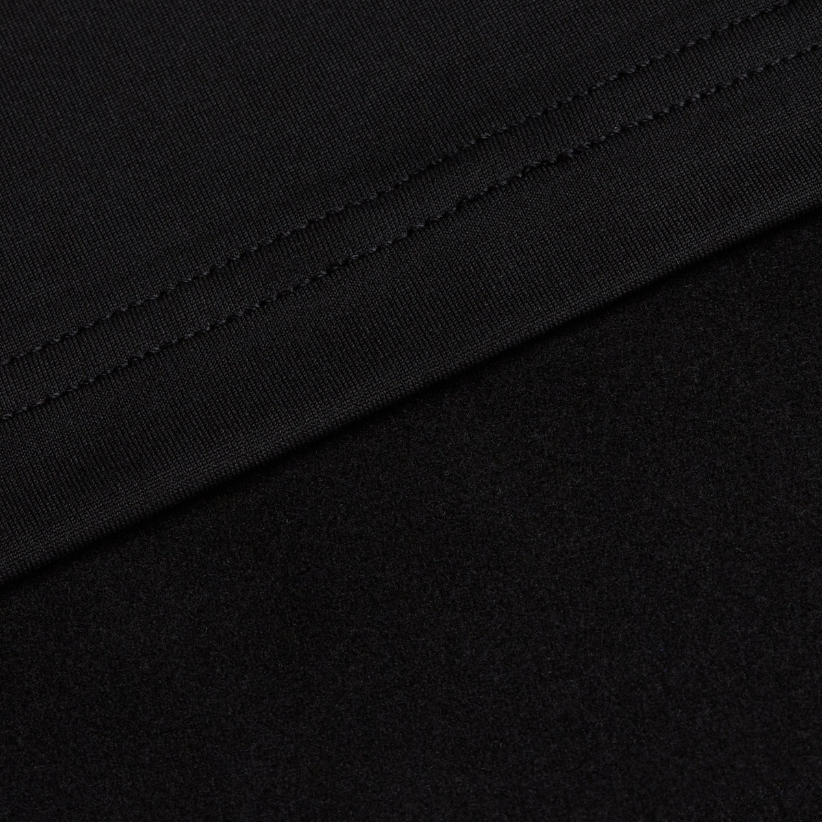 A close-up shot of the Black - Hoodie fabric with a visible seam. The upper section features a tightly woven, smooth textile characteristic of hoodies, while the lower portion reveals a softer, more textured surface. The seam runs diagonally near the top of the image.