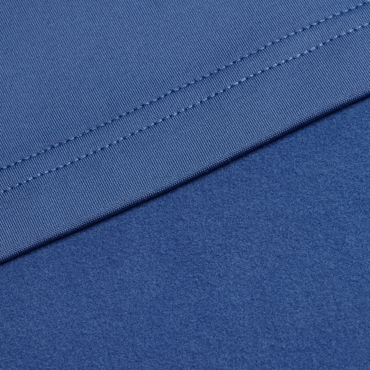 Close-up of blue fabric from the Cooler Blue - Hoodie, showing a seam with parallel stitches running along the top edge. The upper section is smooth and finely woven, while the lower section—characteristic of moisture-wicking materials—displays a slightly textured, matte appearance. Both sections feature similar shades of medium blue, typical for athletic apparel designed for comfort and performance.