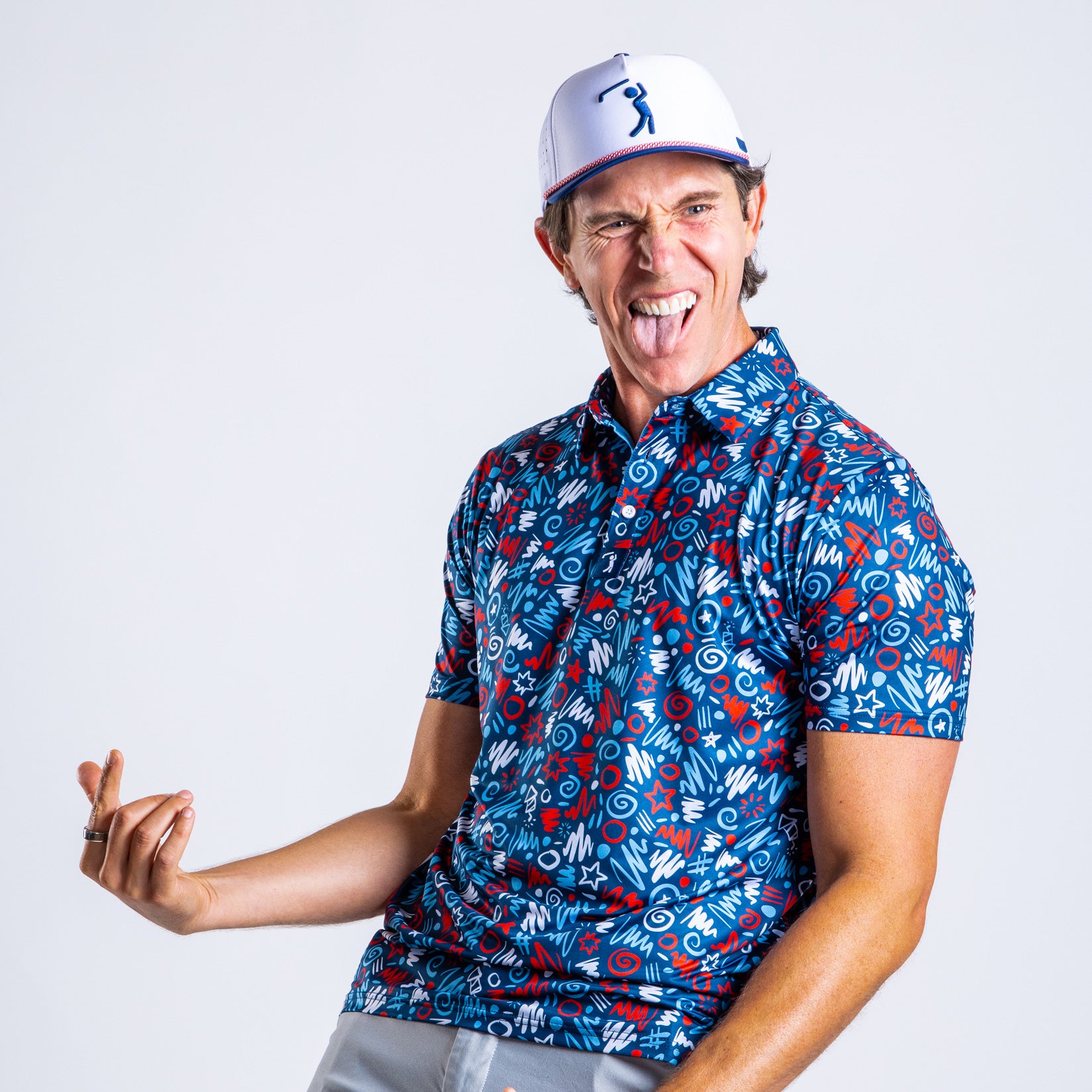 A man in a blue patterned polo shirt and white cap poses playfully, sticking out his tongue and miming an air guitar. He smiles with wide eyes against a plain white background. His outfit features a bold design with red and white shapes.