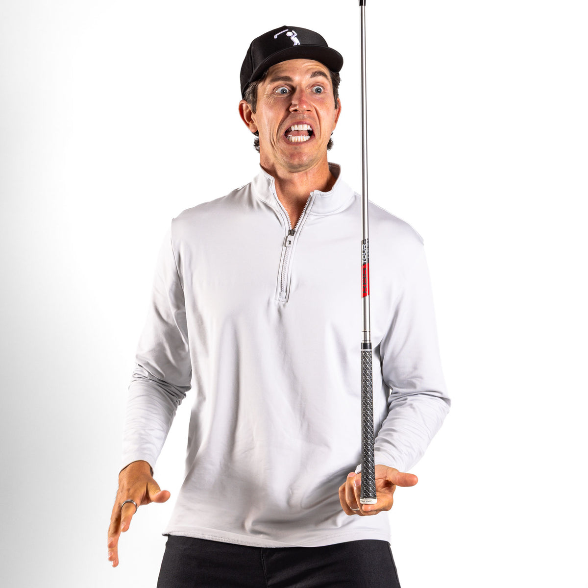 A man wearing the Bone - Q-Zip, a white athletic-fit pullover, and a black cap makes a surprised face while balancing a golf club upright on one finger, standing against a plain white background. His eyes are wide and his mouth is open in excitement.