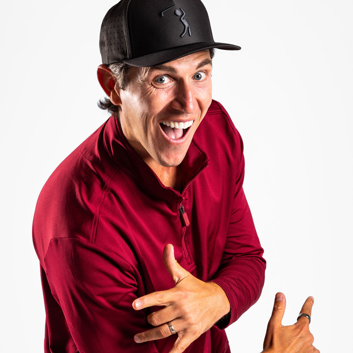 A man with a wide, excited grin wears a black baseball cap with a golf-themed logo and the Burgundy - Q-Zip, a deep red, long-sleeved quarter-zip pullover featuring an athletic fit. He poses playfully against a plain white background, gesturing energetically with his hands while looking directly at the camera.