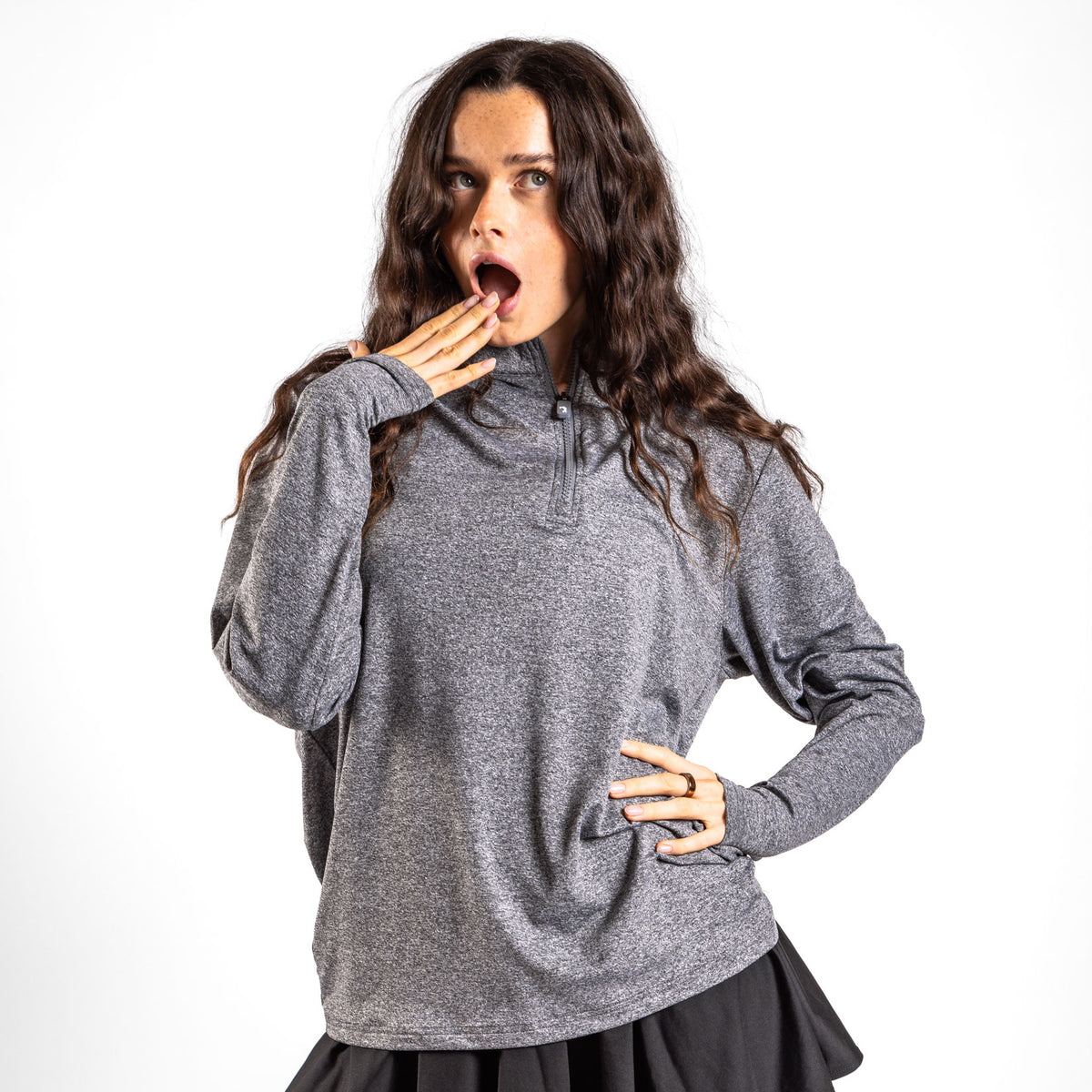 A woman with long wavy hair stands against a white background, looking surprised with her mouth open and one hand to her lips while her other hand rests on her hip. She is wearing the Heather Gray - Q-Zip, which features an athletic fit, paired with a black skirt.