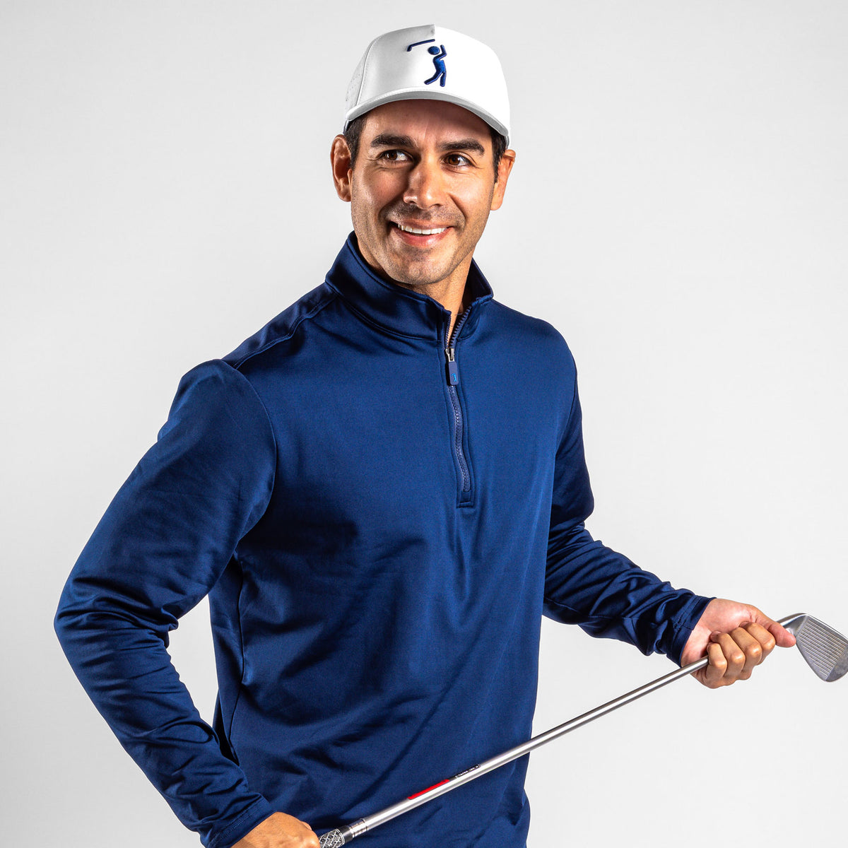 A smiling man with a tan complexion is wearing the Navy - Q-Zip, a navy blue long-sleeved quarter-zip shirt made from moisture-wicking fabric. He pairs it with a white baseball cap featuring a blue golfer logo and looks off to the side while holding a golf iron against a plain light gray background.