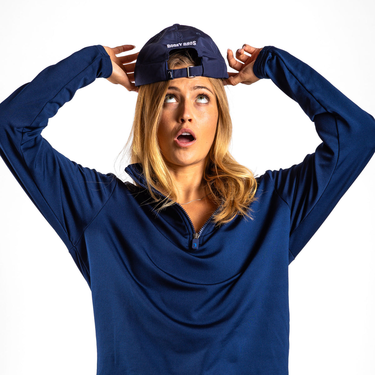 A woman with blonde hair stands against a plain white background, looking up with her mouth open as she adjusts a backward navy cap. She is wearing the Navy Q-Zip, a moisture-wicking pullover.