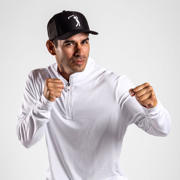 A man in a Black Cap featuring a simple white graphic of a swinging golfer and a White - Q-Zip with an athletic fit poses with fists raised as if boxing, exuding confidence and focus with a slight smirk. The plain light gray background emphasizes his stance. The White - Q-Zip provides UPF 50 protection, making it ideal for active days both indoors and outdoors.