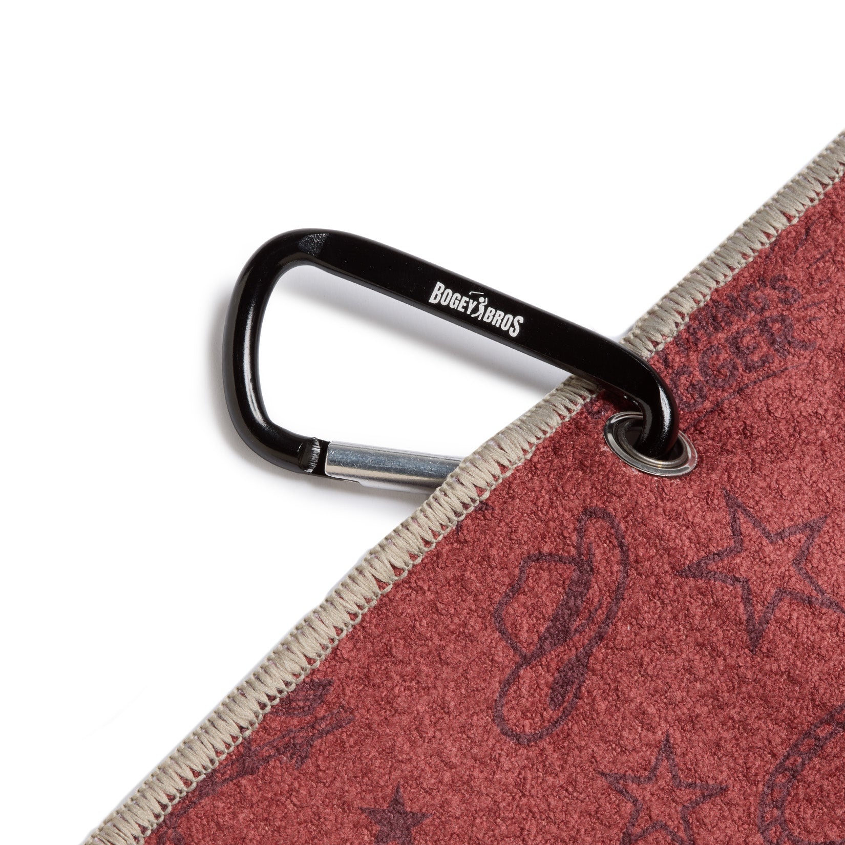 A black carabiner labeled BOGEY BROS clips through a metal grommet on a maroon fabric with western-themed patterns, including stars and cowboy hats. The fabric edge is stitched with beige thread.