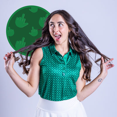 A smiling young woman with long brown hair wears the Birdie Finger - Irish Goodbye Women's Polo, an athletic fit sleeveless green patterned top, paired with a white skirt. She poses playfully against a light gray background with green circle and hand gesture motif.