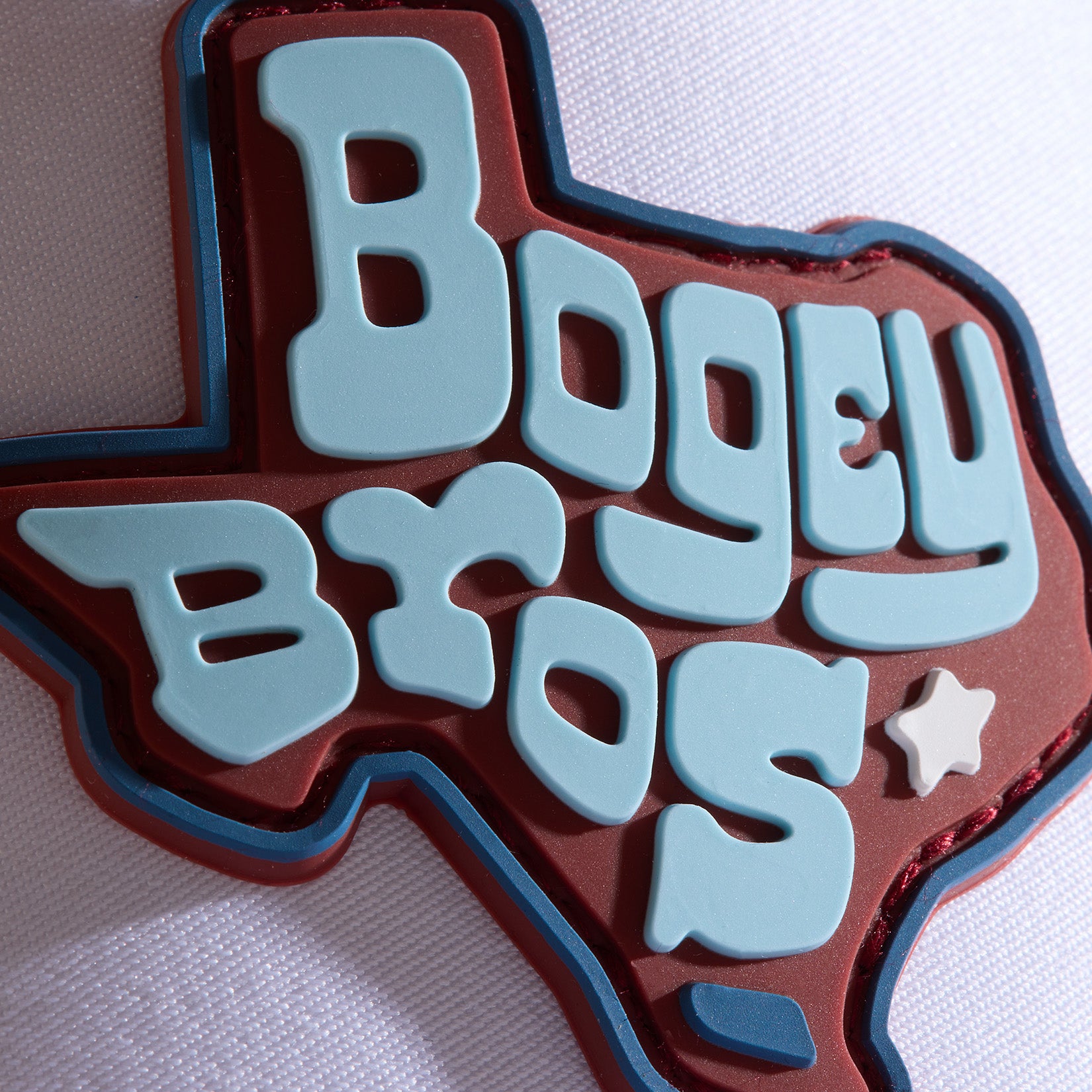 A thick pin shaped like Texas features the words BOGEY BROS in large, light blue letters on a red background, with a small white star under the S. The pin sits on textured white fabric. Total characters: 191.