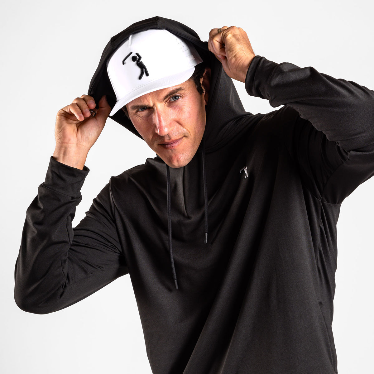 A man wearing the Black - Hoodie pulls the hood over his head, gazing directly at the camera. He pairs it with a white baseball cap featuring a black golfer logo and athletic shorts crafted for comfort and performance. The scene is set against a plain, light grey studio background, highlighting his intense and focused expression.