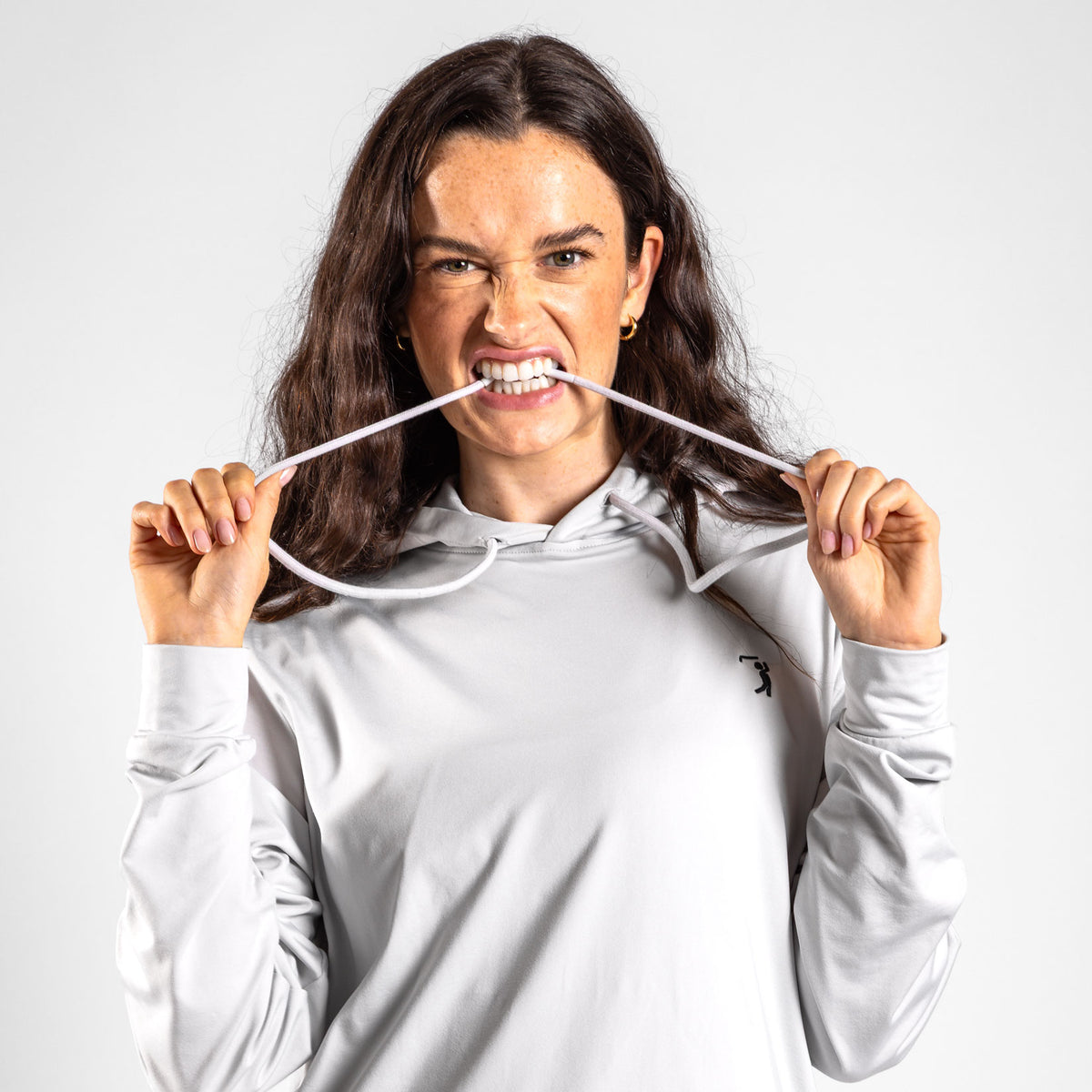 A woman with long brown hair playfully bites and stretches the drawstring of the Bone - Hoodie in light gray, looking directly at the camera with a mischievous expression. The Bone - Hoodie features moisture-wicking fabric to keep her comfortable, set against a plain white background.