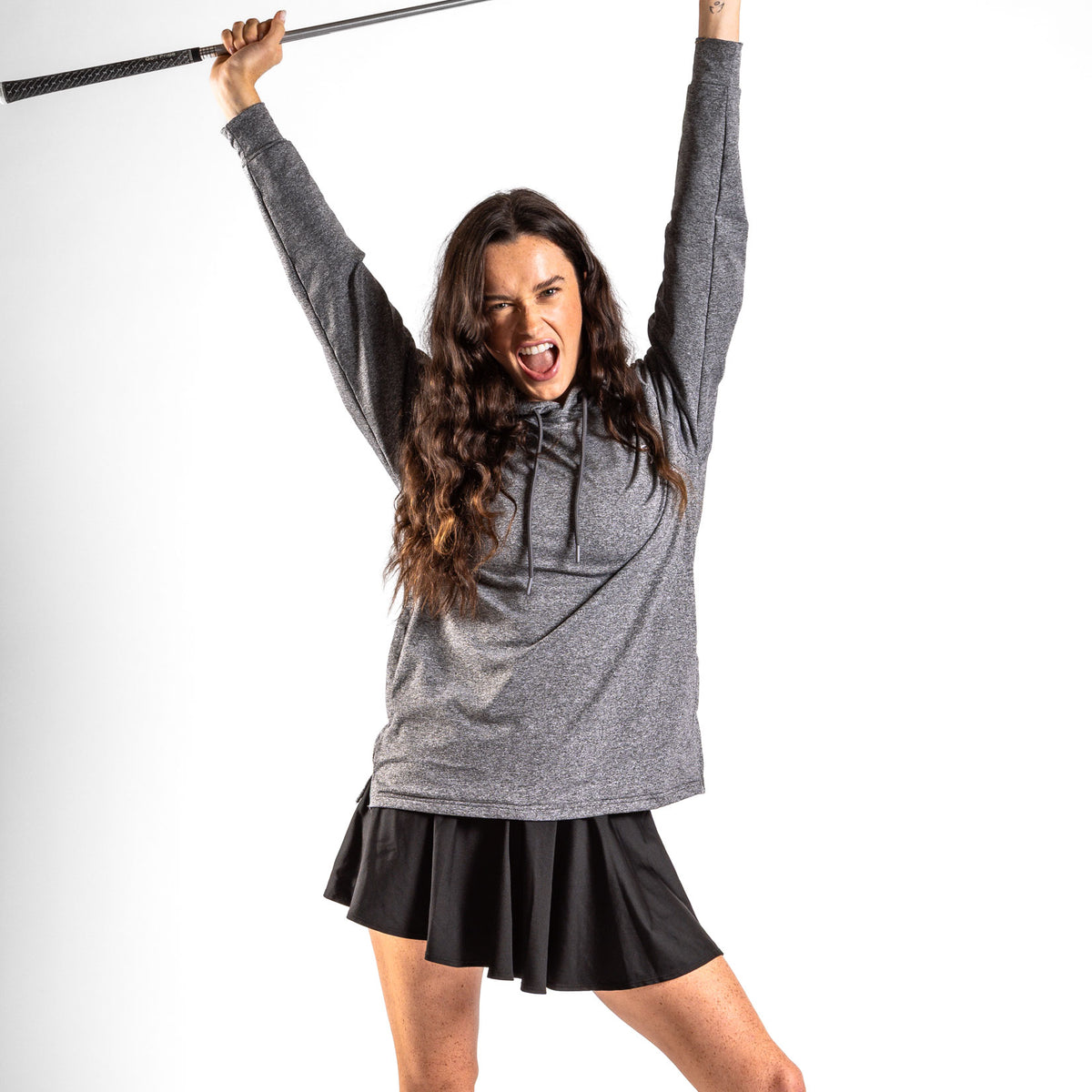 A woman with long dark hair smiles widely, raising a golf club above her head in triumph. She wears the Heather Gray Hoodie, which features moisture-wicking fabric, along with a short black skirt in an athletic fit. The plain white background creates a high-contrast, energetic, and celebratory scene.