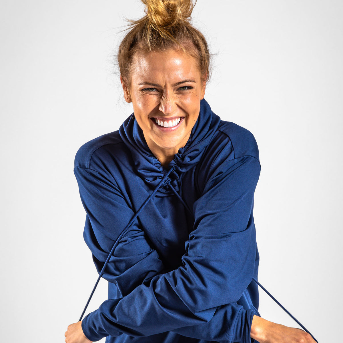 A woman with blond hair in a topknot smiles broadly, her eyes crinkling, as she poses in the Navy - Hoodie made from moisture-wicking fabric. With a playful gesture, she pulls on the hoodie’s drawstrings and crosses her arms in front of her against a plain light gray background.
