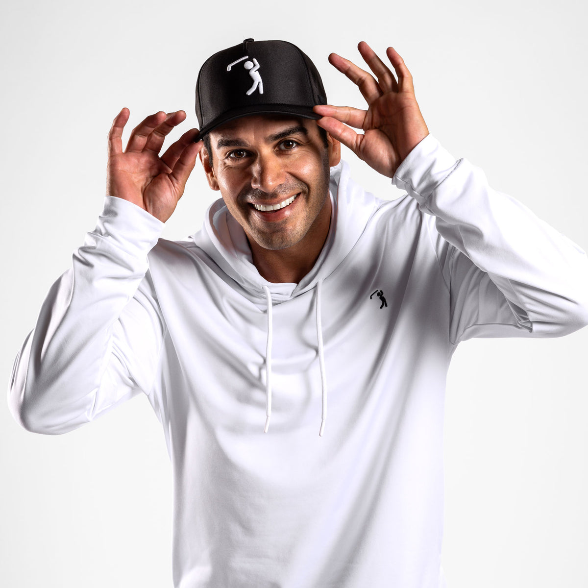 A smiling man wearing the White - Hoodie, which features an athletic fit and a subtle golf swing logo on the chest, adjusts a black baseball cap adorned with a matching logo. The hoodie delivers moisture-wicking comfort, while the plain white background highlights his sporty style.