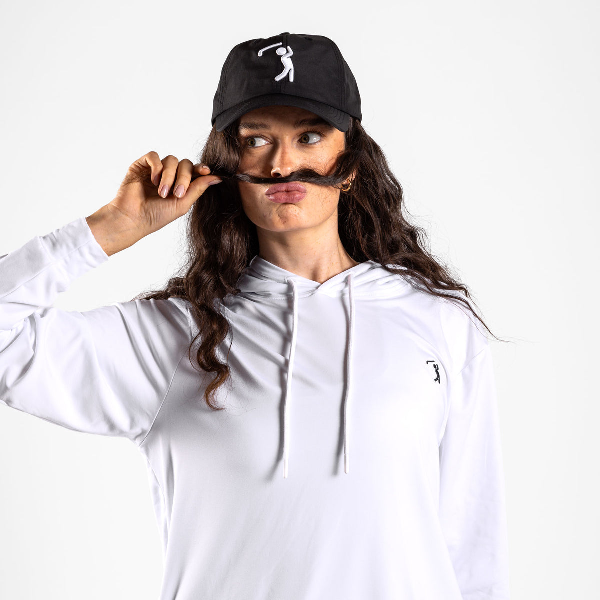 A woman wearing the White - Hoodie and a black cap poses playfully, holding her long brown hair over her upper lip like a mustache and making a pout. The cap, which offers UPF 50 protection, is adorned with a white stick figure graphic. The background is plain white.