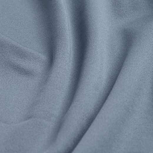 Close-up of matte, dust-blue fabric with a fine, knit-like texture. The material is draped, creating gentle, sweeping folds that transition between highlights and soft shadows, emphasizing the smooth, flexible quality of the textile.