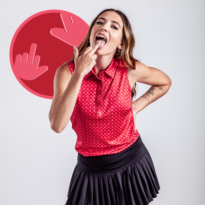 A woman wears the Birdie Finger - Robin - Women's Polo, a sleeveless red patterned athletic top, paired with a black pleated skirt. She poses playfully before a plain backdrop and a bold pink circle graphic showing illustrated rude hand gestures.