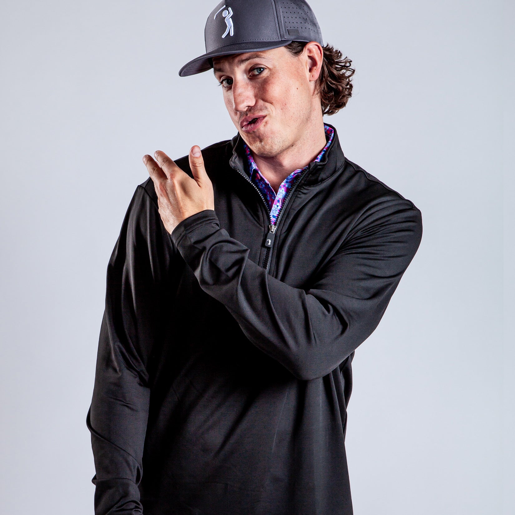 A man in a black zip-up shirt and gray cap gestures playfully with his hand, tilting his head and pursing his lips against a plain light background.