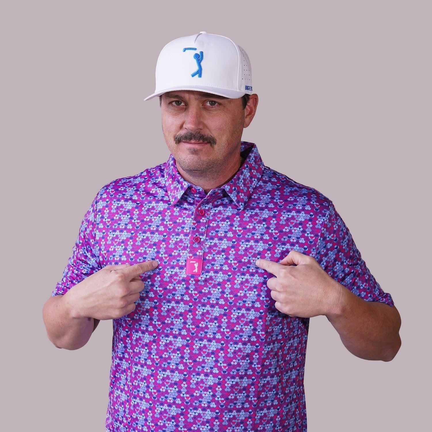 A man with a mustache in a pink and purple patterned shirt and white cap points at a pink logo on his shirt, standing against a plain light background.