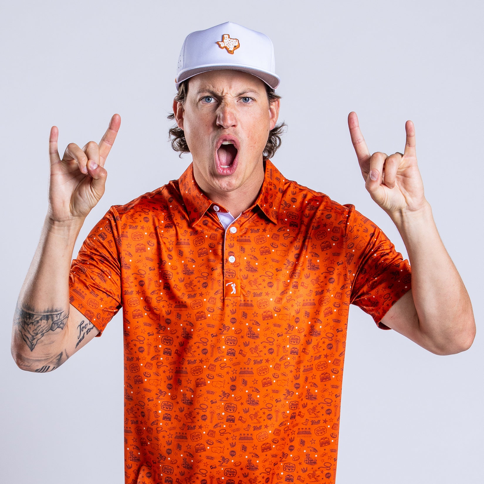 A man in an orange patterned shirt and white cap making a “hook em horns” gesture with both hands, mouth open as if shouting, standing against a plain light background. His hat has an orange Texas outline logo, and he has a tattoo on his left forearm.