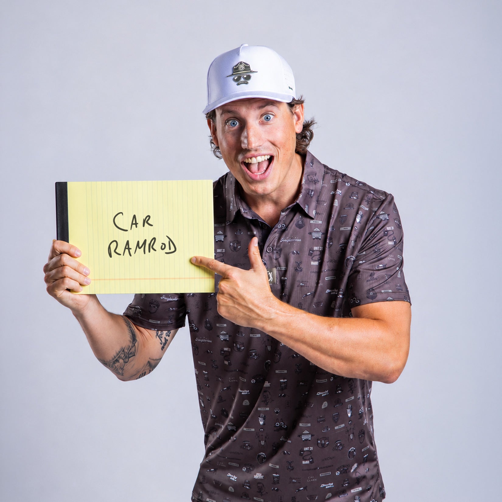 A smiling man in a printed shirt and white cap holds a yellow notepad that reads CAR RAMROD in black marker. He points at the text with his right hand, standing against a plain light grey background. His expression is enthusiastic and playful.