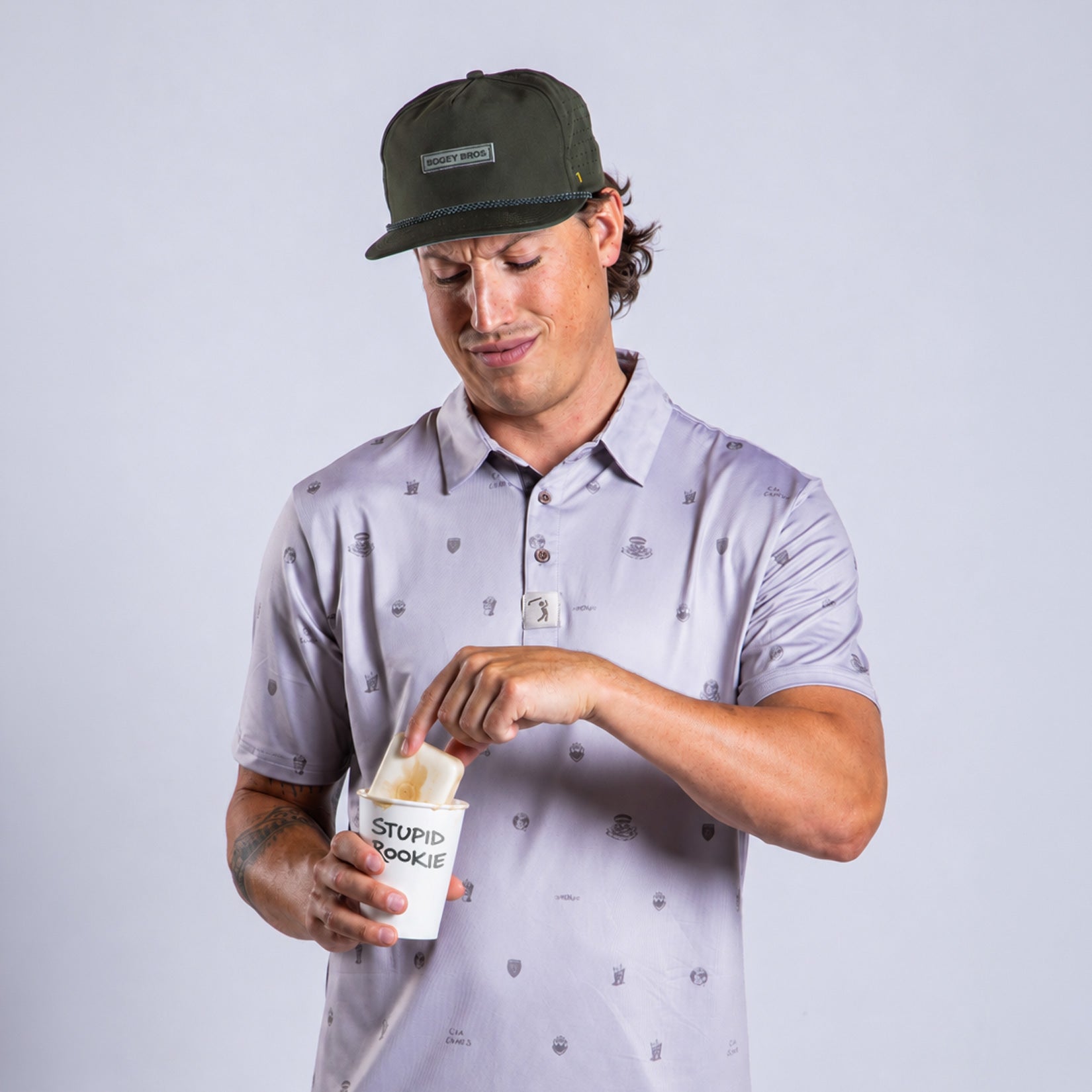 A man in a gray patterned polo and green cap looks down, pulling a cookie from a white cup labeled STUPID COOKIE with one hand. He has a slight smile and stands against a plain light gray background.