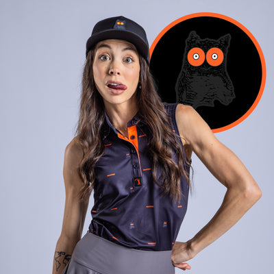 A woman with long brown hair, wearing a black hat and the Night Owl - Women's Polo with an orange and black owl logo, sticks out her tongue playfully. The athletic-fit polo provides UPF 50 protection and has a bold owl logo on the back.