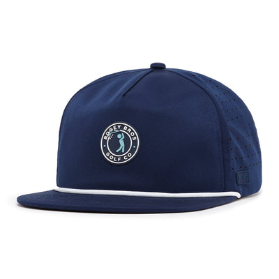 The Bogeyman Stamp Navy Unstructured Flat Bill Hat is a navy snapback featuring white accent piping, an embroidered circular “Bogey Bros Golf Co” golfer logo, side ventilation holes, and performance material construction.