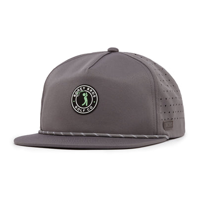 The Bogeyman Stamp Charcoal is an unstructured flat bill snapback hat with a round black and white Casey Bros Golf Co. logo patch, performance fabric, side ventilation holes, and a thin braided rope across the front.