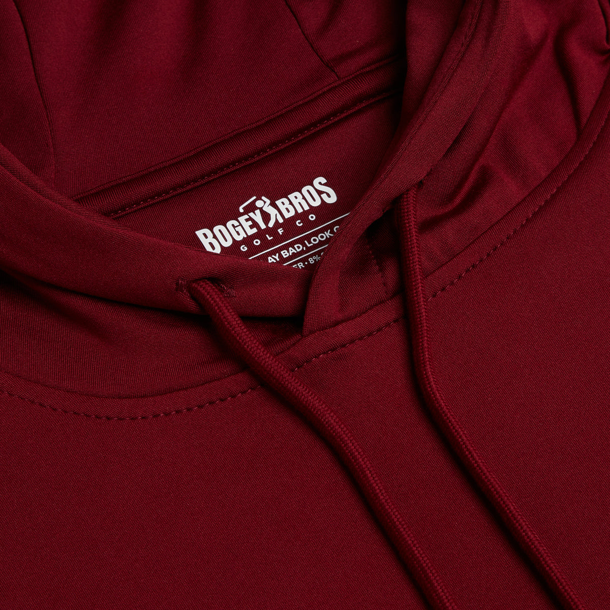 A close-up of the Burgundy - Hoodie showcases a hood with drawstrings and an inner label that reads Bogey Bros Golf Co. MY BAD, LOOK GOOD. The soft, smooth fabric exhibits neat stitching along the edges and features moisture-wicking properties to ensure all-day comfort.