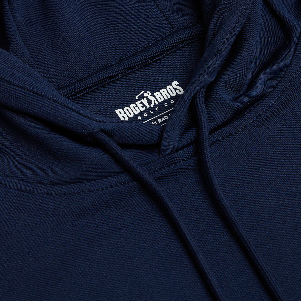 A close-up, top-down view of the Navy - Hoodie showcases its dark navy blue color and thoughtful design. The interior neck label is visible, displaying crisp white text that reads BOGEY BROS GOLF CO alongside a golf icon, and a partial line beneath it says AY BAD. Crafted for active comfort, this hoodie features moisture-wicking fabric to help keep you dry whether you’re on or off the course. Matching navy drawstrings hang from the neckline of the hood.