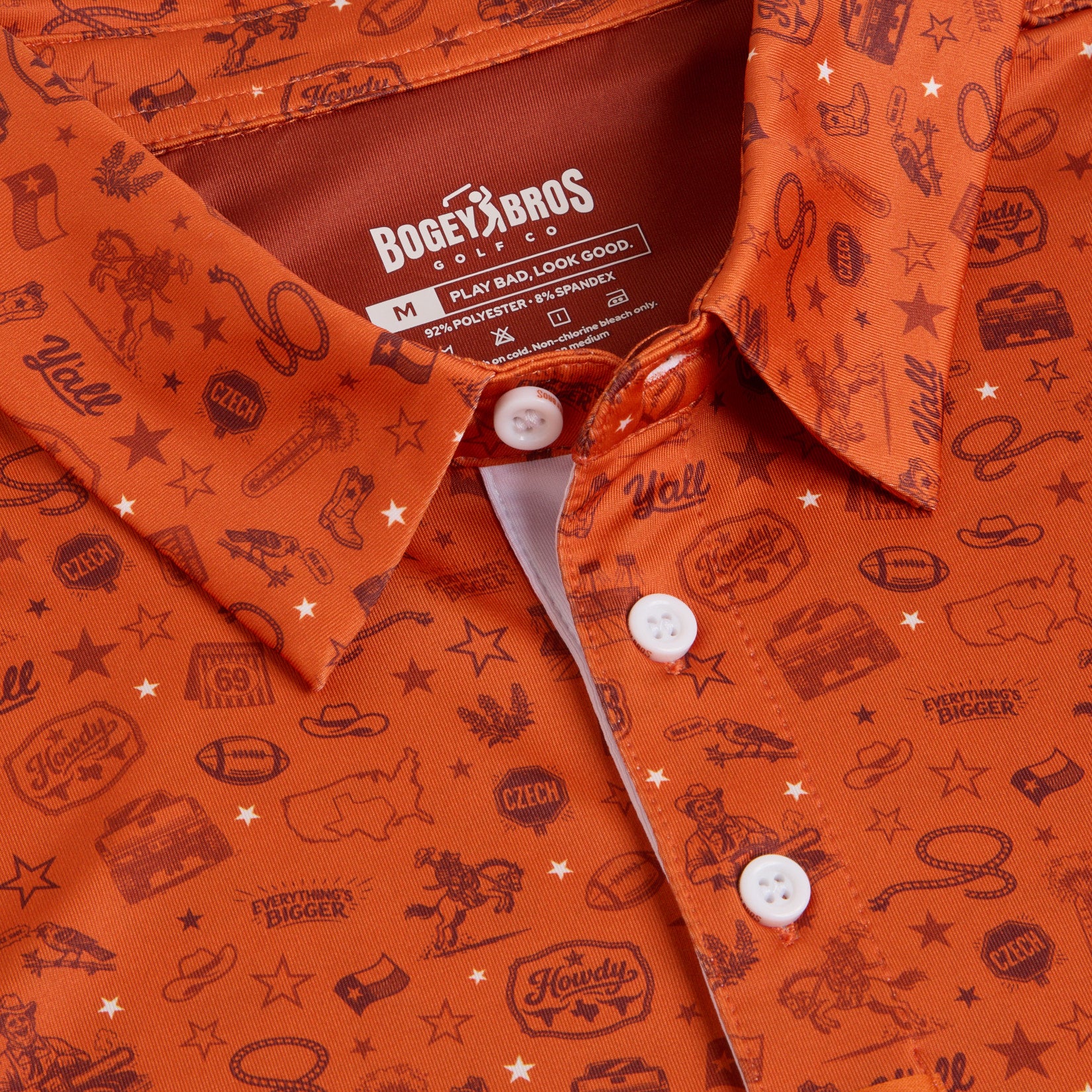 Close-up of an orange shirt with white buttons. The fabric displays dark orange Western motifs, including boots, hats, guitars, stars, cowboy figures, lassos, and text like Howdy and Yall. The label reads “Bogey Bros” and gives sizing and care instructions.