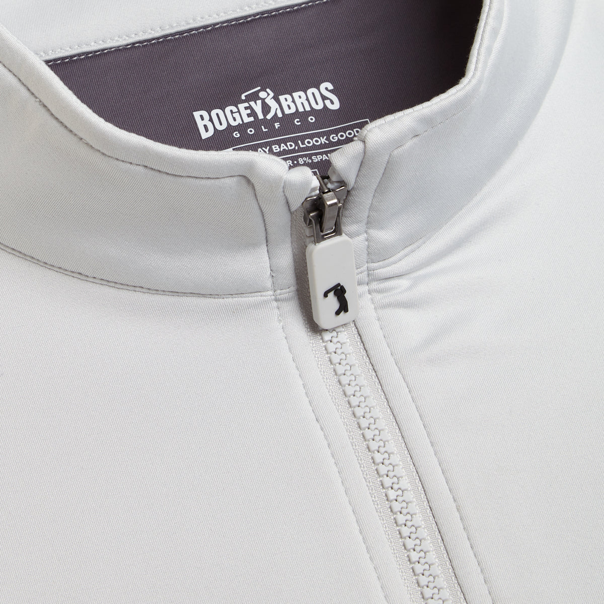 Close-up of the Bone - Q-Zip’s upper section reveals a vertical zipper with a white pull adorned by a black golf club graphic. The inside label displays “Bogey Bros Golf Co.” in white text against dark fabric. It features a rounded, soft collar and an athletic fit designed to deliver both comfort and style for any activity.