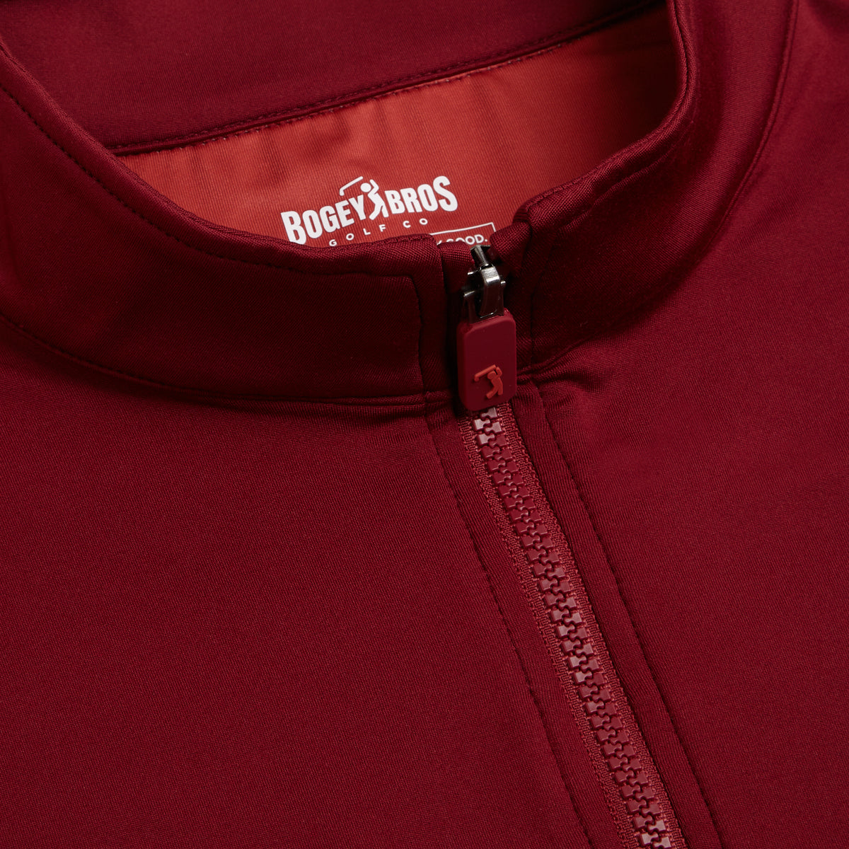 Close-up of the Burgundy - Q-Zip, partially zipped. The zipper pull features a small golfer icon, and inside the collar, BOGEY BROS GOLF CO. is printed in white uppercase letters on a maroon fabric background. Crafted with an athletic fit and moisture-wicking fabric, this jacket delivers comfort and performance both on and off the course.