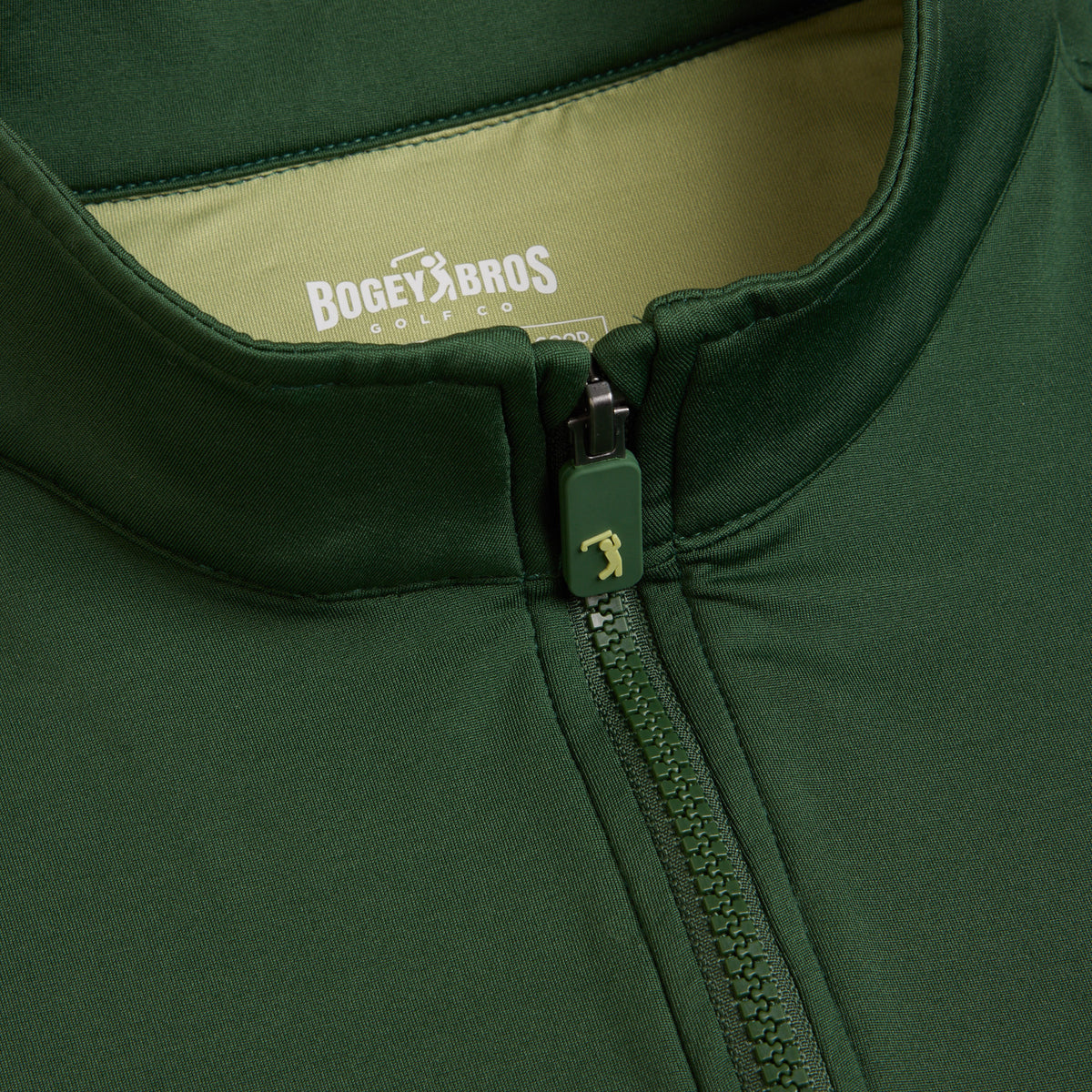 Close-up of the Forest Green - Q-Zip, a dark green zip-up jacket with a high collar tailored for an athletic fit. The zipper pull is embellished with a golfer logo, and the inside lining is light green. The label displays “BOGEY BROS GOLF CO.” in white letters beneath a small golfing figure.