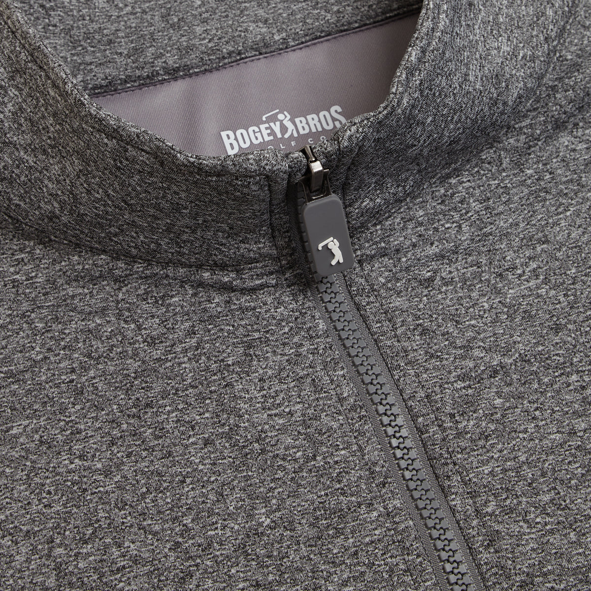 Close-up of the Heather Gray - Q-Zip jacket. The zipper is partially up, displaying a small golf club logo on the pull. Inside the collar, a gray label shows the brand name BOGEY BROS in white text. Made with textured, soft-looking fabric, this jacket offers an athletic fit and moisture-wicking properties for all-day comfort.