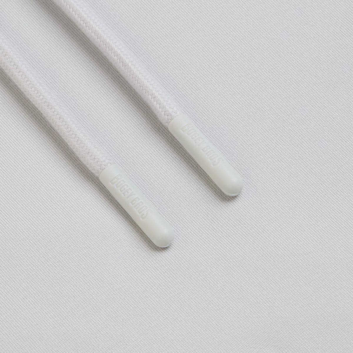 Close-up of two white shoelace tips, known as aglets, lying parallel on a white textured fabric. The aglets are made of plastic and have the word AGLET embossed on them. Reminiscent of the clean design featured in the Bone - Hoodie, which is crafted with moisture-wicking properties and a minimalist aesthetic, the overall image conveys a sense of simplicity and freshness.