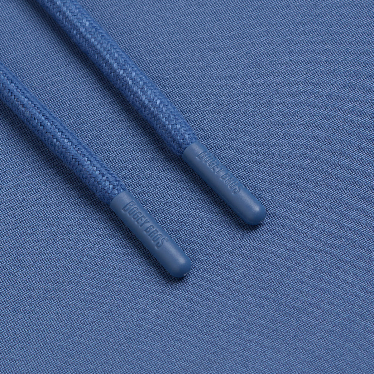 Two blue drawstring tips rest on matching blue fabric from the Cooler Blue - Hoodie, reminiscent of the high-performance material found in moisture-wicking athletic shorts. The cylindrical tips are made of smooth plastic, each embossed with LACE LINKS. The drawstrings have a zigzag pattern while the hoodie fabric features a fine texture.