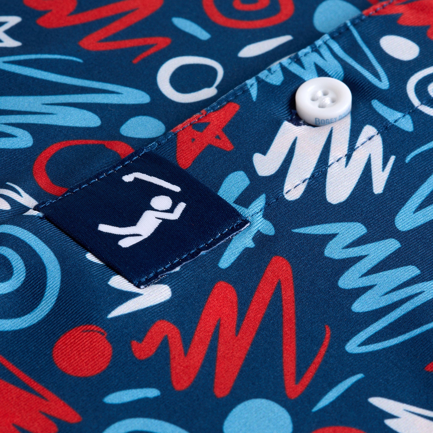 A close-up of a fabric with a vibrant, abstract pattern in red, blue, light blue, and white. The design features swirls, zigzags, circles, and lines. A white button and a navy tag with a white minimalist human figure are also visible.