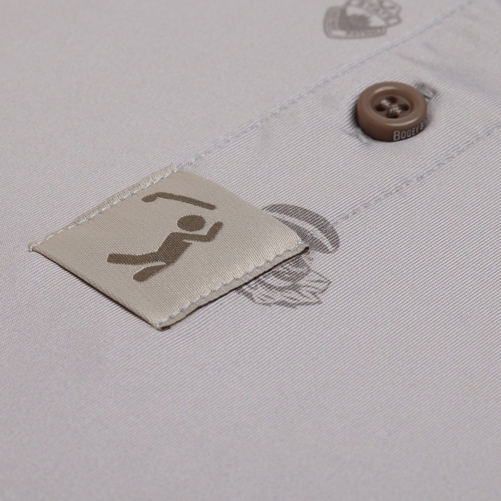 A close-up of a beige shirt shows a brown button and a stitched tag with a simple dark brown icon of a person lifting weights. The shirt fabric has a smooth texture, and faint printed graphics are partially visible in the background.