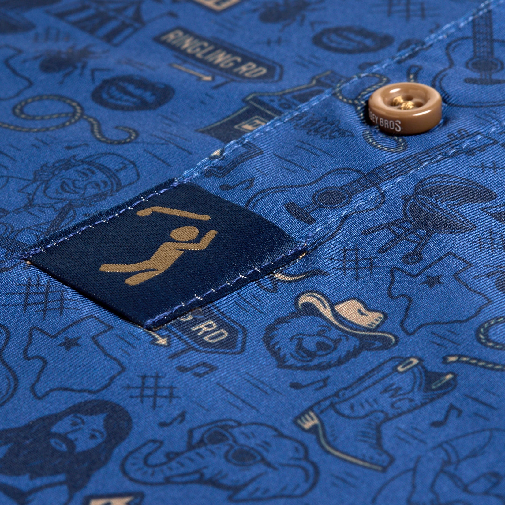 Close-up of the Ringling Road - Polo, a blue shirt adorned with brown buttons and playful line art featuring cowboy hats, guitars, boots, musical notes, faces, a microphone, and a gold tag displaying a pictogram of a person with drumsticks. The fabric showcases western and musical motifs throughout.