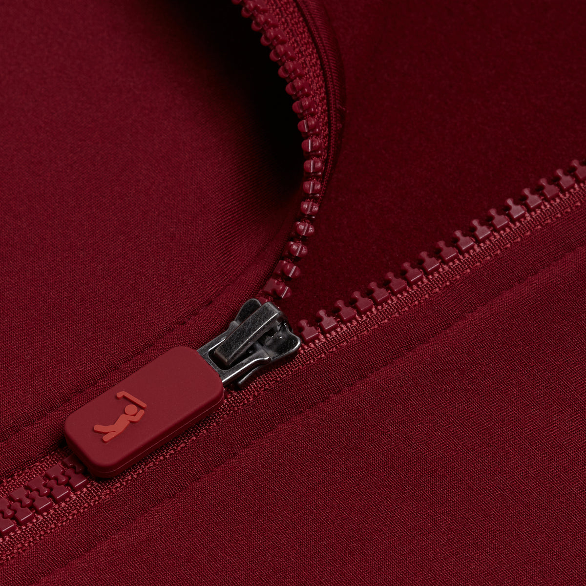 Close-up of a partially unzipped Burgundy - Q-Zip on matching red fabric. The zipper pull features a small raised logo, while the texture of the moisture-wicking material and the plastic zipper teeth are clearly visible.