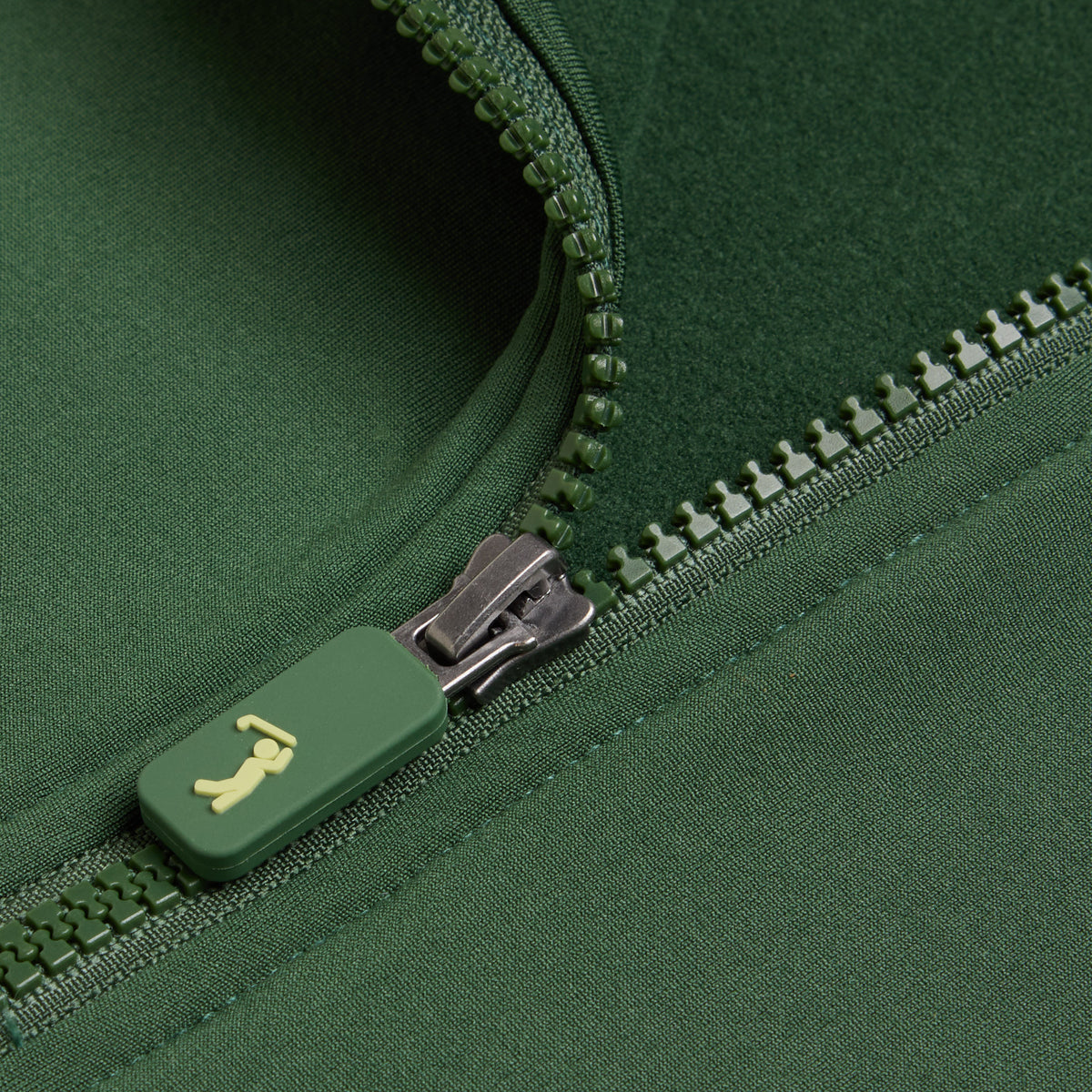 A close-up of the Forest Green - Q-Zip showcases an athletic-fit garment with a green zipper. The matching green zipper pull features a small yellow, stylized animal logo, possibly resembling a dog. The surrounding fabric is smooth, moisture-wicking, and consistent in its rich forest green hue to keep you comfortable during activities.