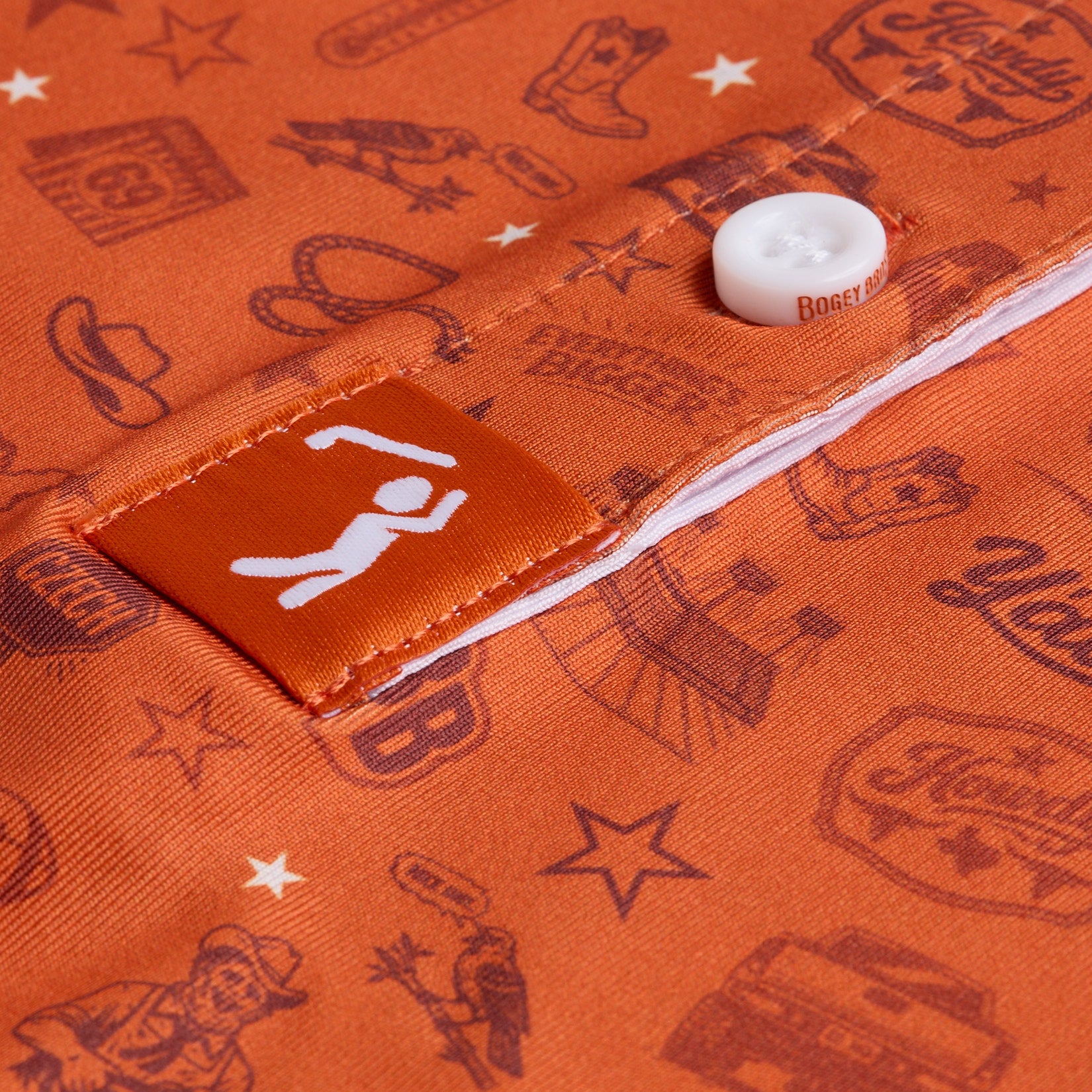 Close-up of orange fabric with western-themed illustrations in dark red, featuring stars, cowboy hats, boots, and faces. A white button is sewn on a pocket flap, and a small orange tag with a white icon of a golfer raising arms is stitched nearby.