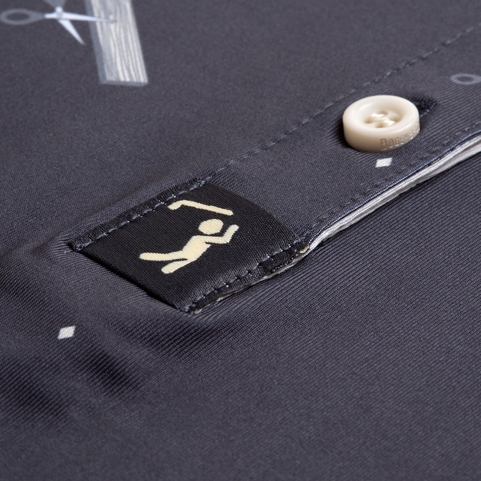 Close-up of the Scissor Me Timbers - Women's Polo in dark gray, featuring a cream button and a black tag with a climber and ice axe. The smooth fabric has subtle geometric patterns and provides UPF 50 sun protection for outdoor adventures.