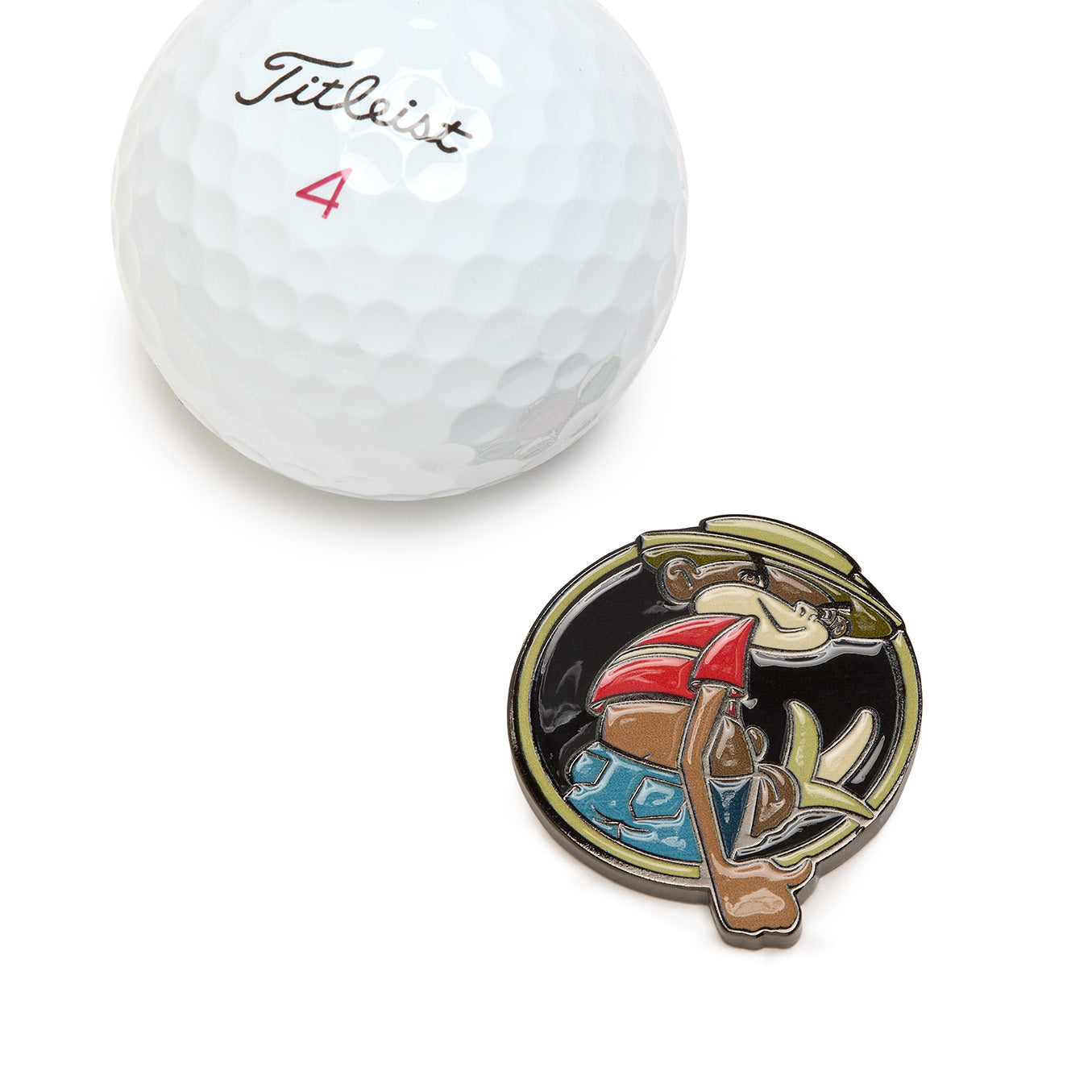 A white Titleist 4 golf ball sits beside the Johnny Chimpo - Ball Marker, a heavy metal accessory with iron nickel plating, featuring a circular pin decorated with a cartoon monkey in a hat, sunglasses, blue shorts, and red shirt giving a thumbs up—all set against a black background.