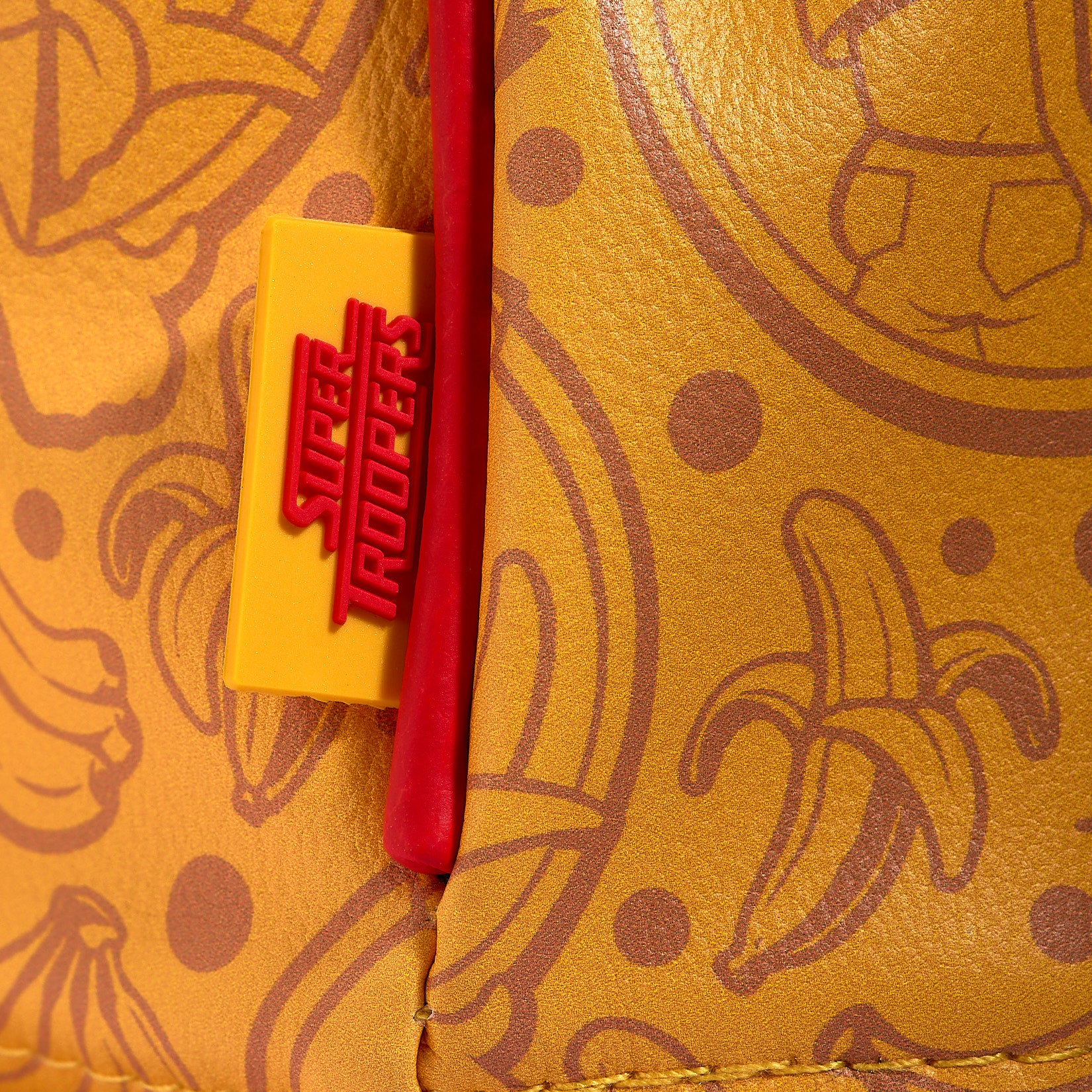 Close-up of the Johnny Chimpo - Driver Headcover, showcasing yellow material with brown banana and dot patterns, a yellow woven golf accessory tab labeled SUPER TROOPERS in red, and a thick red cord overlapping the tab. The texture suggests premium PU leather or high-quality faux leather construction.