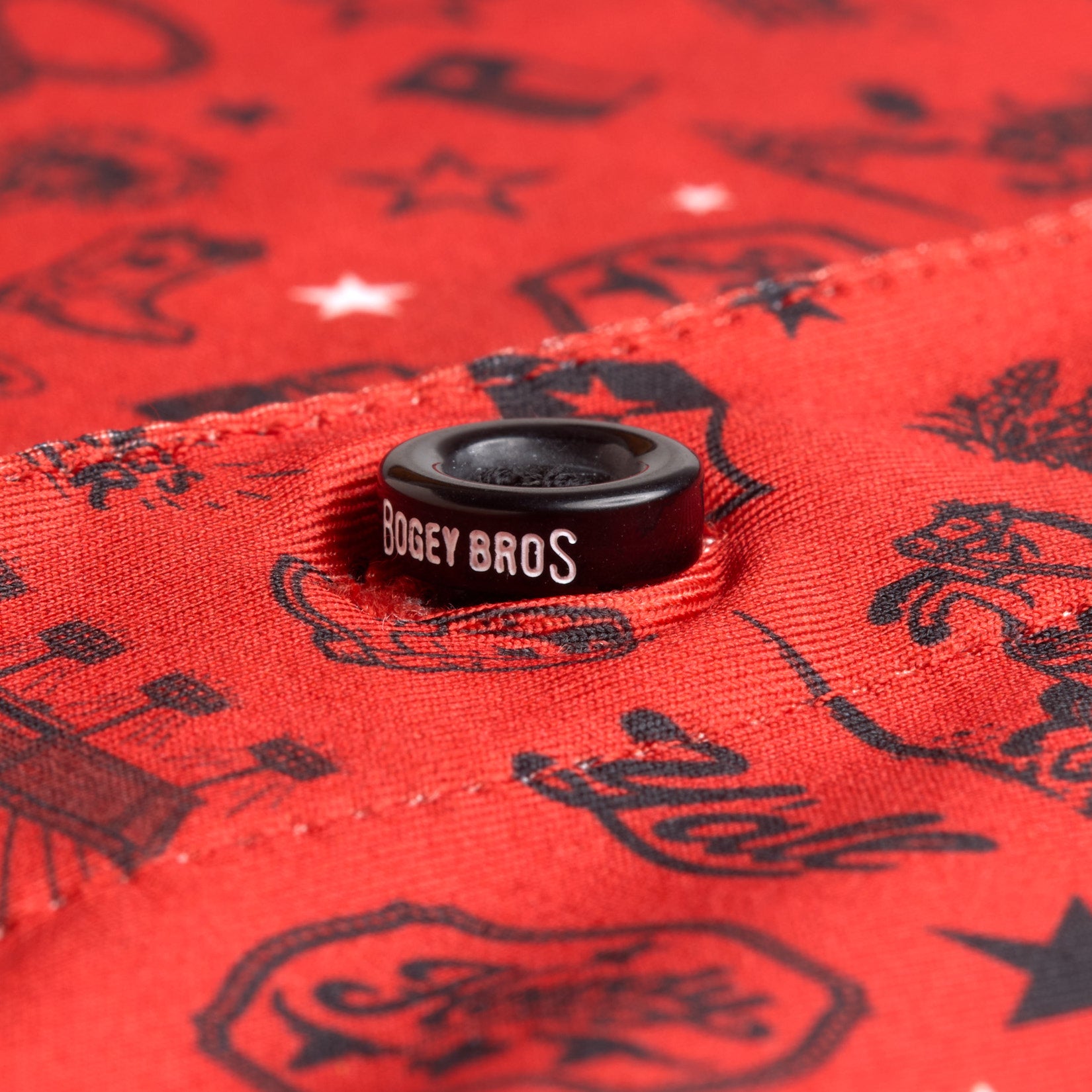 A close-up of a red fabric with black graphic patterns and a black button. The button has BOGEY BROS printed in white letters. The fabric features various small illustrations, including stars, flames, and other abstract shapes.