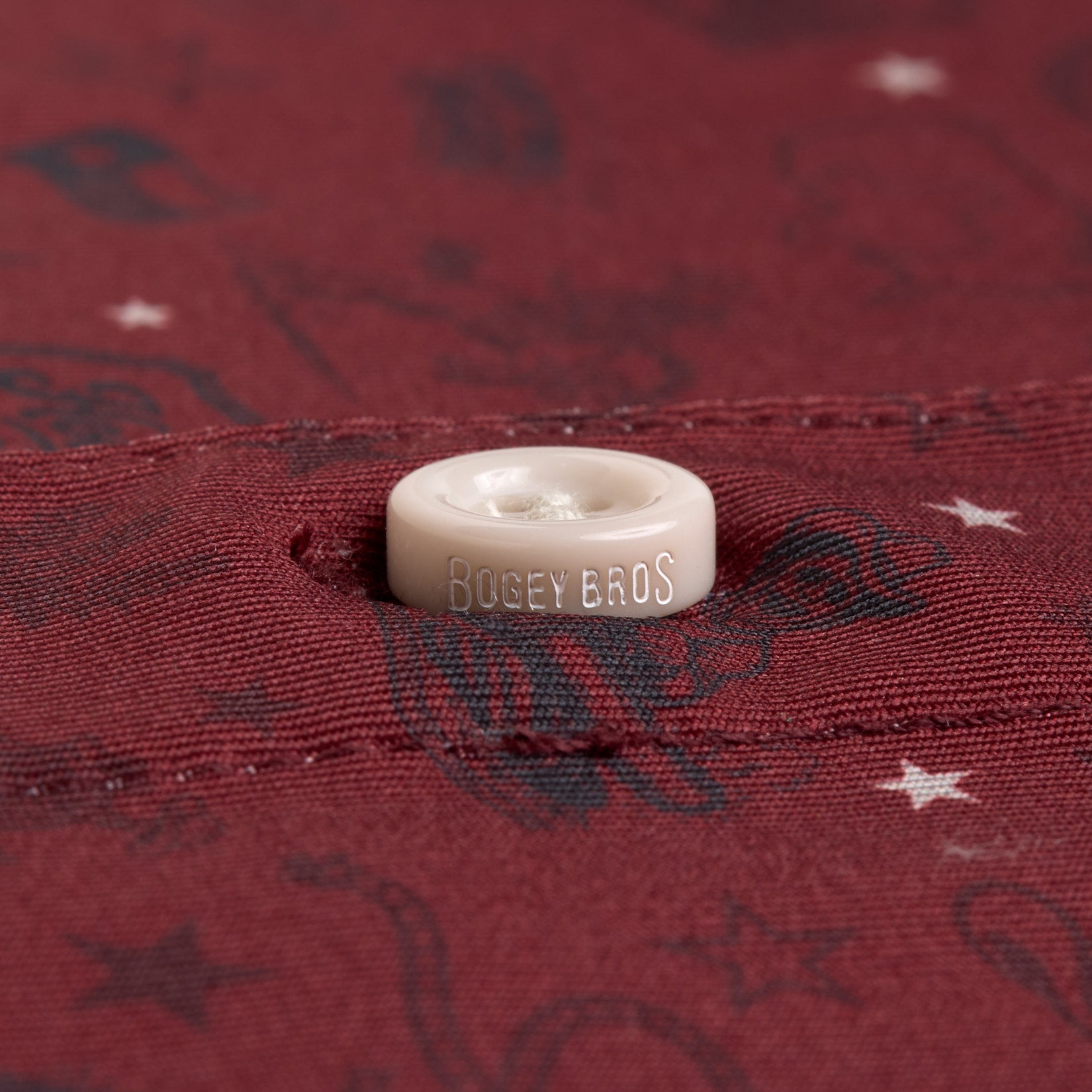 A close-up of a maroon fabric with subtle patterns and white stars. A cream-colored button is sewn onto the fabric, engraved with the words BOGEY BROS. The stitching and buttonhole are visible, emphasizing the detail and texture of the material.