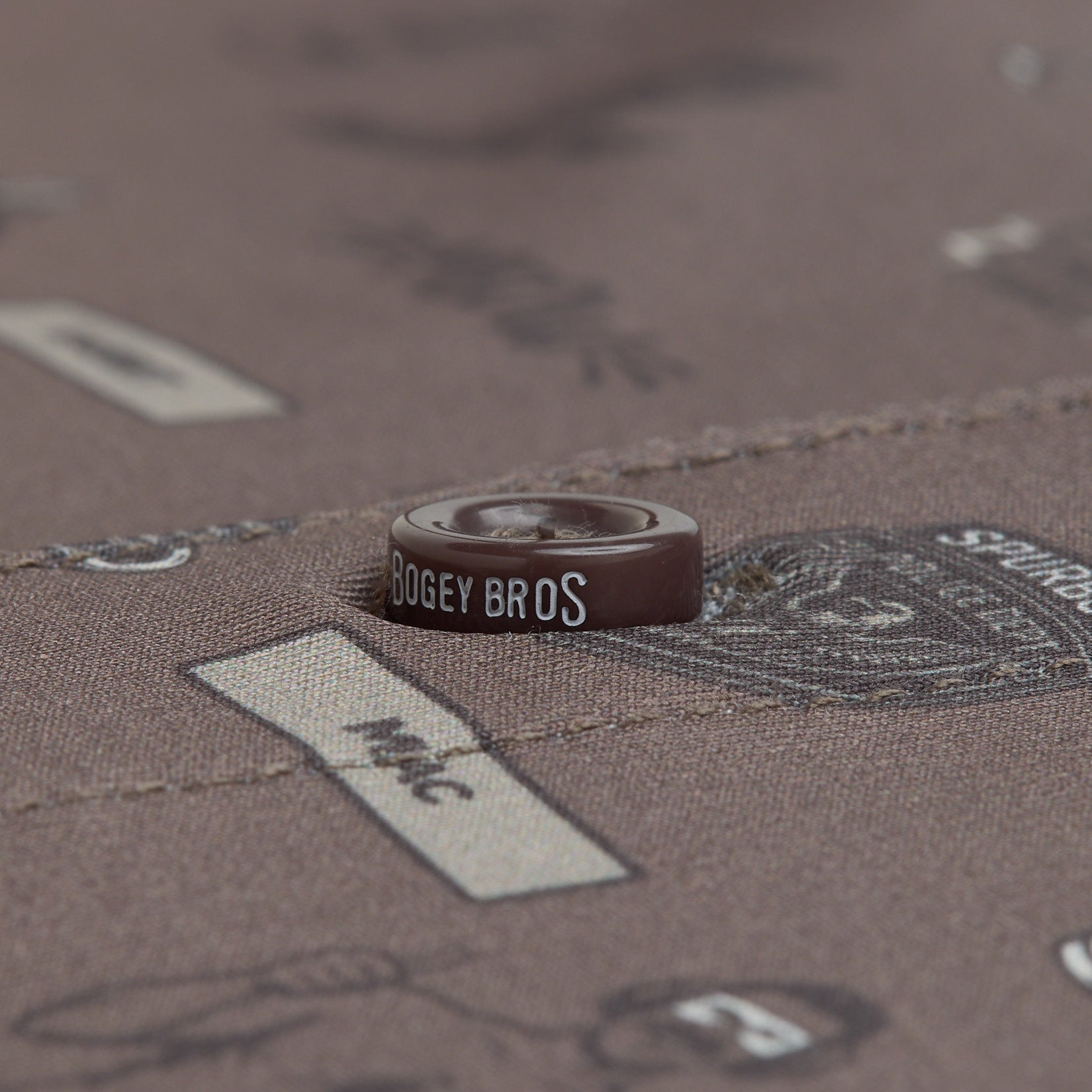 A close-up of a brown button sewn onto brown fabric with printed designs and text. The button has BOGEY BROS written in white letters. The fabric features black line drawings and beige rectangles with various words and images.