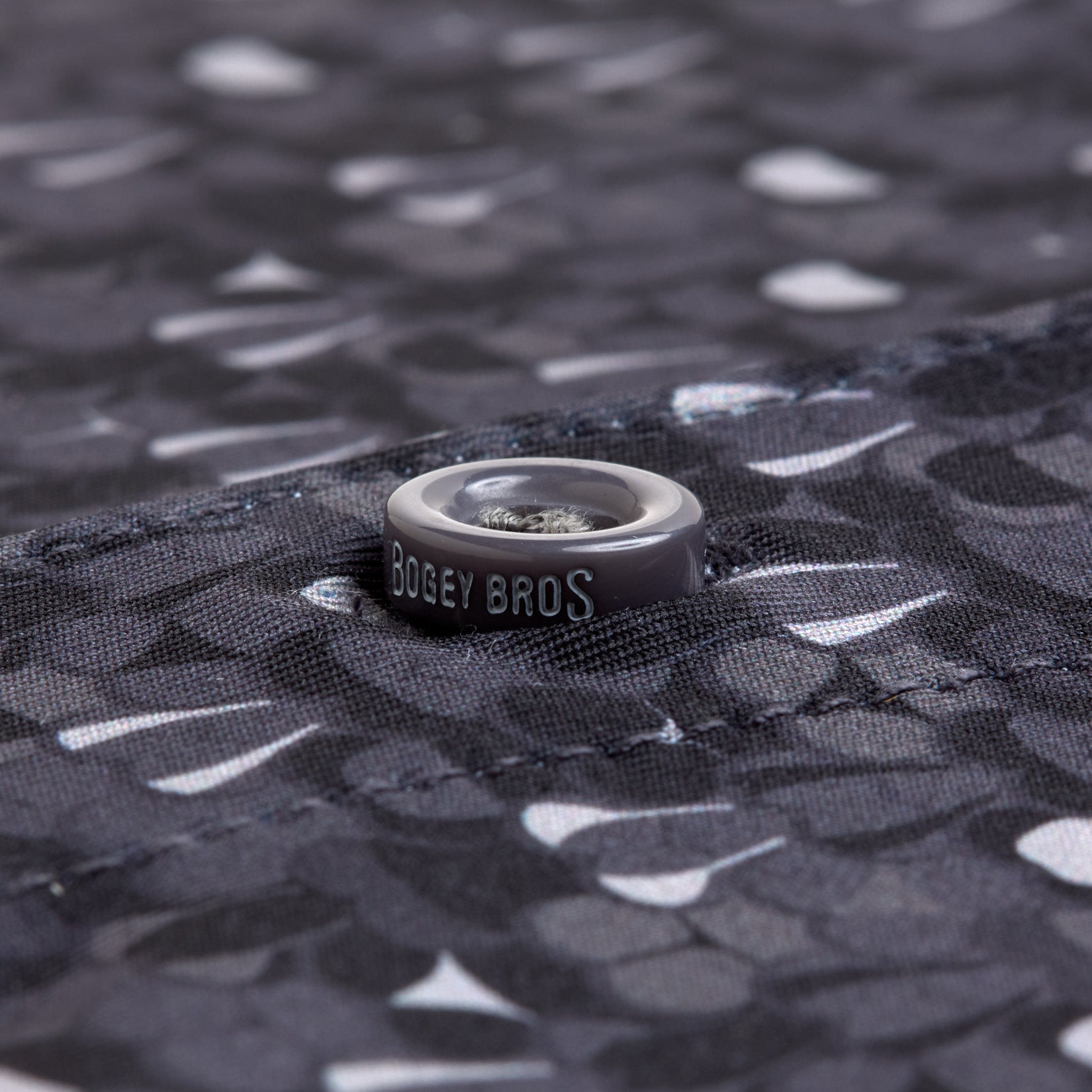 Close-up of a gray button with "BOGEY BROS" printed on a dark patterned fabric featuring white golf tees. The T&A - Fitty Shades - Polo shirt offers moisture-wicking for all-day comfort.